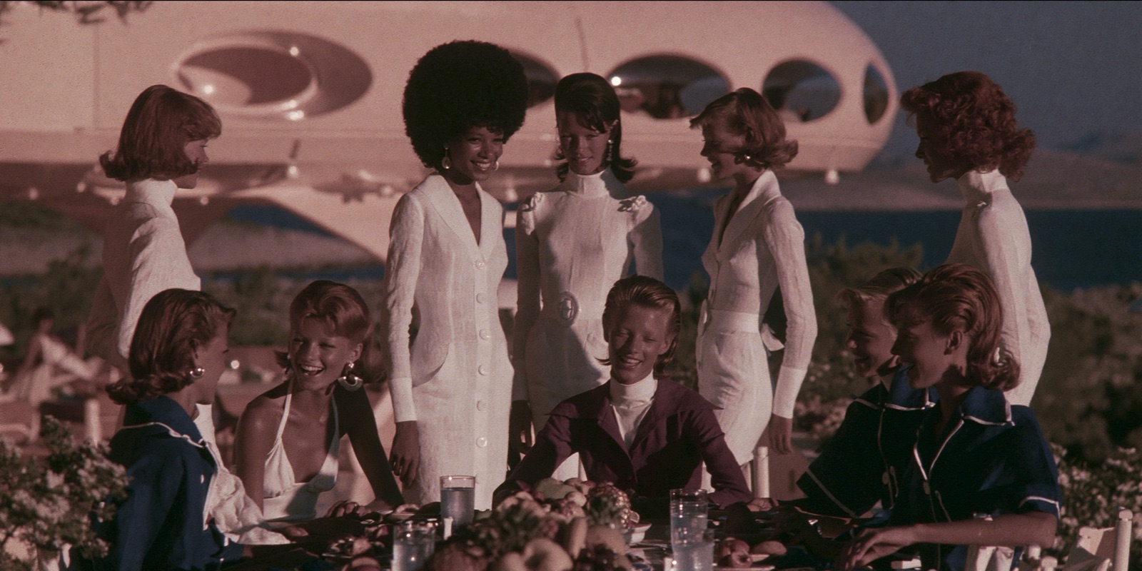 Seven of the crew at a long table above the water, all dressed alike.